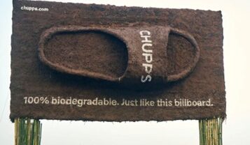 A large, sandal-shaped billboard made of brown, textured material displays the text "100% biodegradable. Just like this billboard."