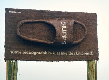 A large, sandal-shaped billboard made of brown, textured material displays the text "100% biodegradable. Just like this billboard."
