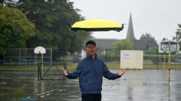 A young man stands beneath a flying yellow umbrella in the rain, with a basketball court and trees in the background.