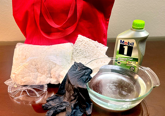 Red tote bag, two packages of material, black gloves, safety goggles, glass bowl, and a bottle of Mobil 1 fuel on a wooden surface.