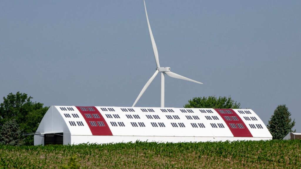 A white barn with red panels and solar windows, beside a tall wind turbine, set against a clear blue sky and green trees.