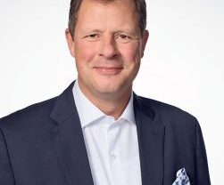 Man in a navy suit and white shirt, smiling, with a patterned pocket square against a white background.