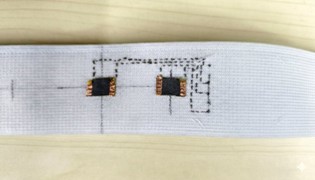 A close-up view of a white fabric strip featuring two small black electronic chips and circuit patterns printed in black.
