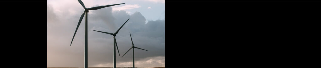 Three wind turbines stand against a cloudy sky, showcasing their blades and structure in a landscape with muted earth tones.