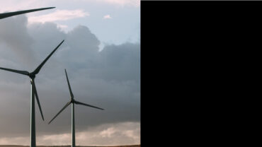 Three wind turbines stand against a cloudy sky, showcasing their blades and structure in a landscape with muted earth tones.
