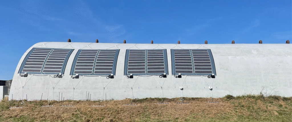 Solar panels mounted on a curved gray structure against a bright blue sky, with grass and supports visible at the bottom.