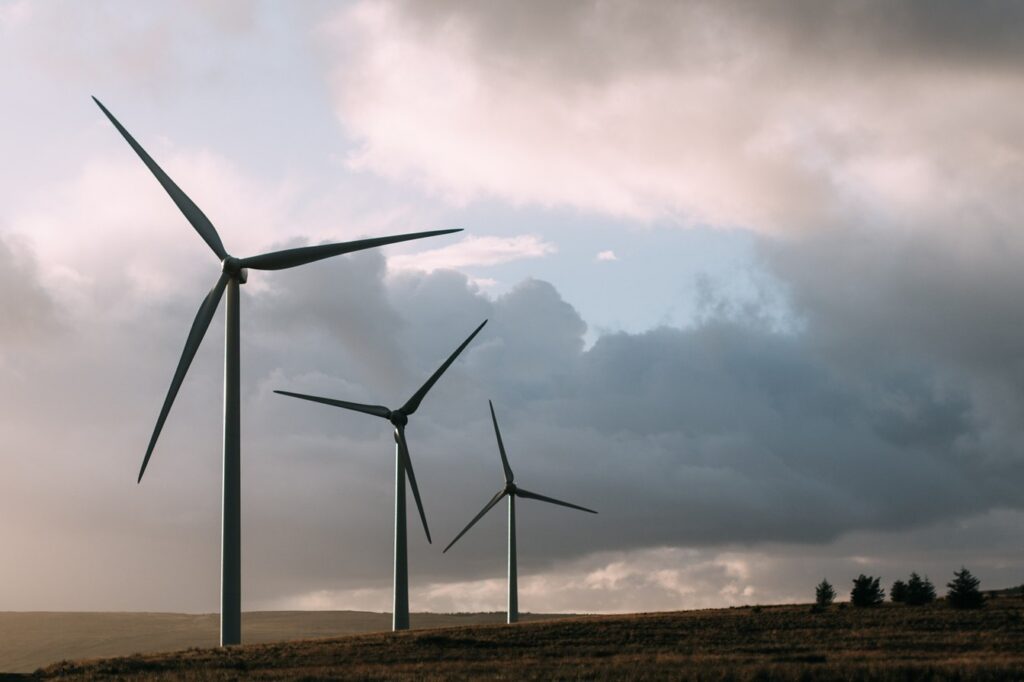 Three tall wind turbines stand against a cloudy sky, with rolling hills beneath them, symbolizing renewable energy and sustainability.
