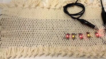 A woven, beige textile with fringed edges displays colored LED lights (red and yellow) connected by a black wire.