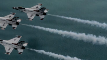 Five fighter jets fly in a tight formation above a blue ocean, leaving white vapor trails against the sky.