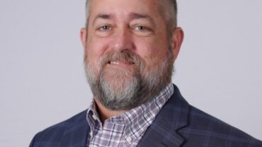 Smiling man with a beard, wearing a blue plaid blazer over a checkered shirt, set against a light gray background.