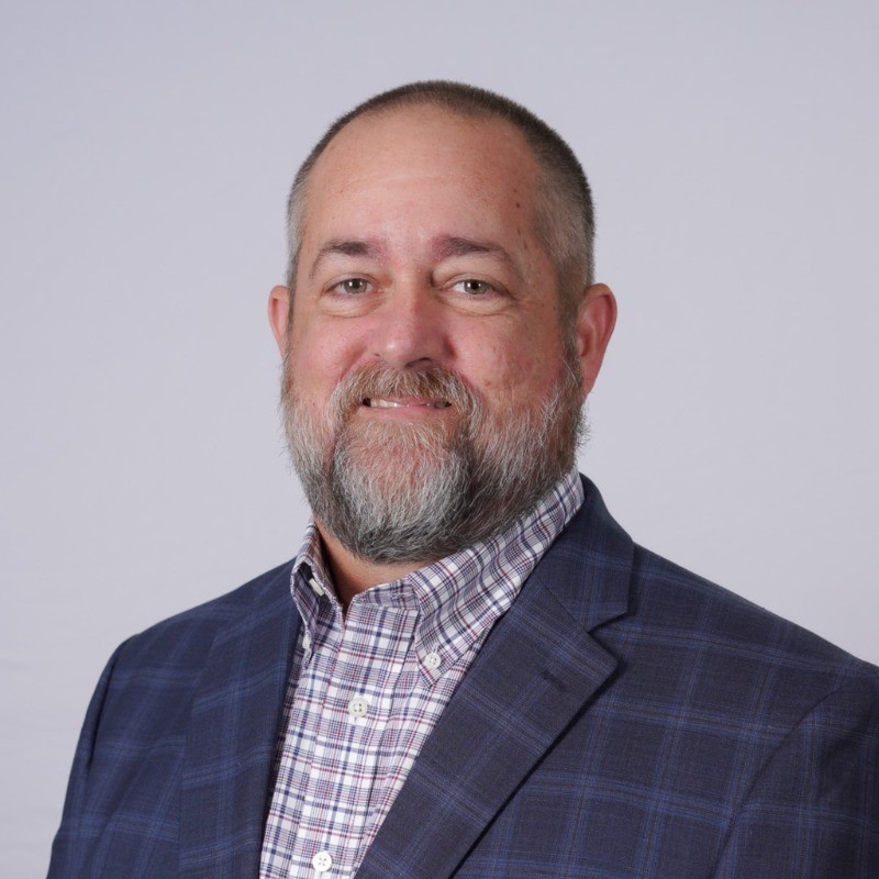 Tony Smiling man with a beard, wearing a blue plaid blazer over a checkered shirt, set against a light gray background.