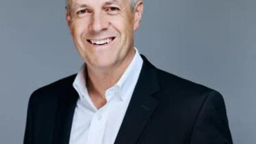 Smiling man in a black suit with a white shirt, set against a neutral gray background.