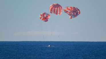 Three red and white parachutes descend over blue ocean water, with a small spacecraft visible below them on the surface.