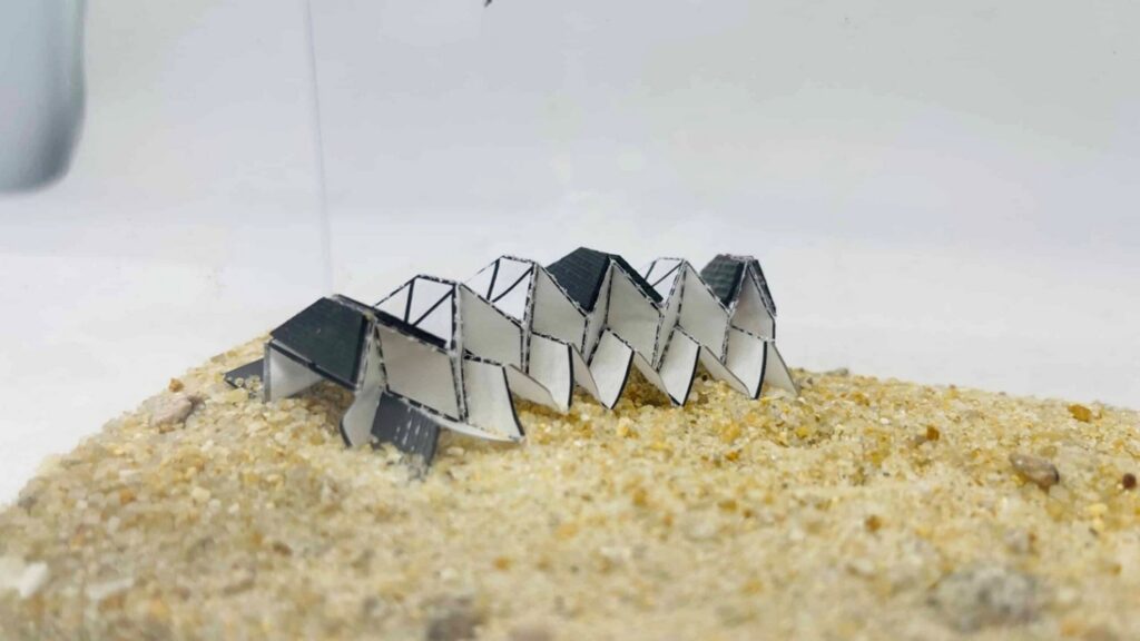 A row of origami-like robotic structures in gray and white, set on a textured sandy surface, showcase innovative biomedical engineering.