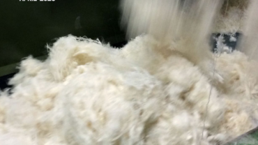 Textured fibers are flowing from a metal chute, with the title "Tipping the Scale" prominently displayed above. The background is green.