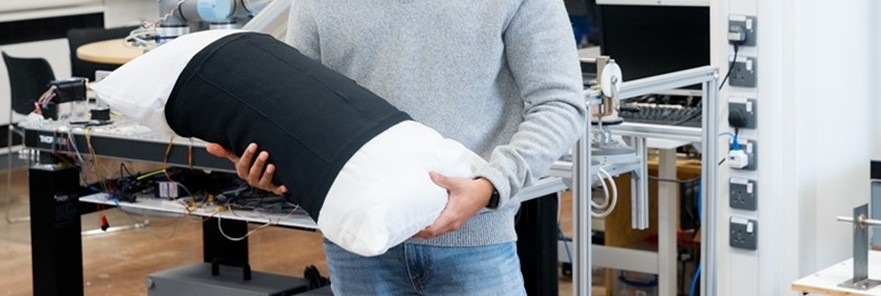 A person holds a smart pillow sleeve with a black and white design, set against a backdrop of a technology lab with machinery and equipment.
