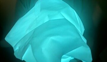 Glowing blue fabric is held in hands, creating a soft light effect against a dark background. The texture appears smooth and crumpled.