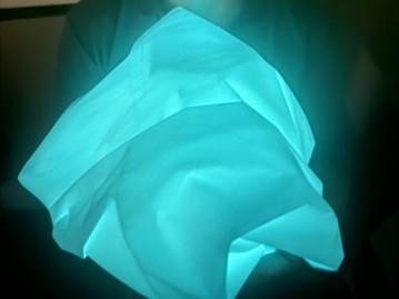 Glowing blue fabric is held in hands, creating a soft light effect against a dark background. The texture appears smooth and crumpled.