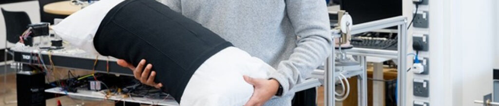 A person in a light sweater holds a large, cylindrical medical textile with a black fabric cover and white padding, against a tech-filled backdrop.