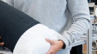 A person in a light sweater holds a large, cylindrical medical textile with a black fabric cover and white padding, against a tech-filled backdrop.