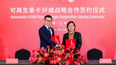 Executives from The LYCRA Company and Texhong International Group shake hands, holding red folders, with a floral arrangement in front.