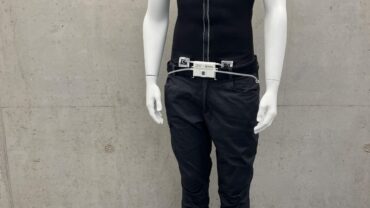 A mannequin displays black protective workwear, including a sleeveless top and pants, alongside worn work boots on a gray floor.