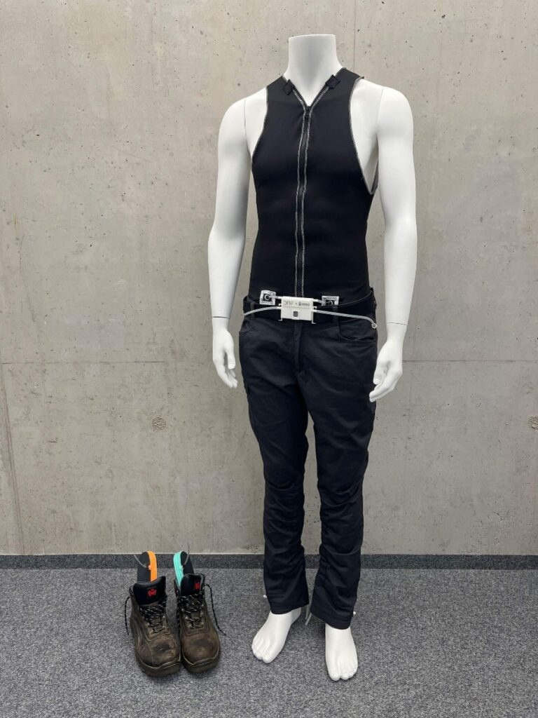 A mannequin displays black protective workwear, including a sleeveless top and pants, alongside worn work boots on a gray floor.
