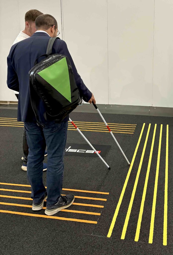 Two individuals stand on a textured floor, one using a white cane, near yellow tactile guidance lines and a black logo on the ground.