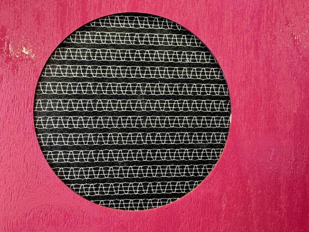 A circular cutout in a bright pink background reveals a black fabric featuring white wavy lines, suggesting a textile design.