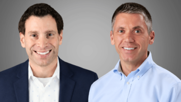 Two smiling men in professional attire, one in a dark blazer with a patterned shirt and the other in a light blue button-down shirt, against a gray background.