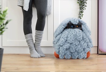 A soft, fluffy blue robot sits beside a person wearing gray leggings and cozy socks, with a light-colored wall and plants in the background.