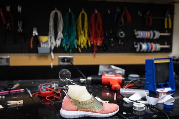 A pink shoe on a cluttered workbench, surrounded by tools and colorful cords hanging on the wall in the background.