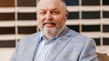 A smiling man with a beard, wearing a light blue jacket and shirt, sits casually in front of a wall displaying various color swatches.