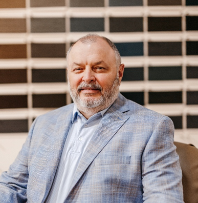 A smiling man with a beard, wearing a light blue jacket and shirt, sits casually in front of a wall displaying various color swatches.