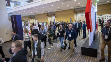 Crowd of attendees walking through a modern convention center, with registration signs and booths visible in the background.