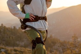 A person wearing a gray and green exoskeleton stands on a rocky terrain, with mountains in the background and a sunset glow.
