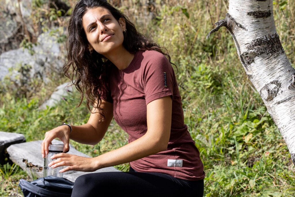 A woman sitting near a birch tree, wearing a maroon shirt and black leggings, holding a water bottle, with greenery in the background.