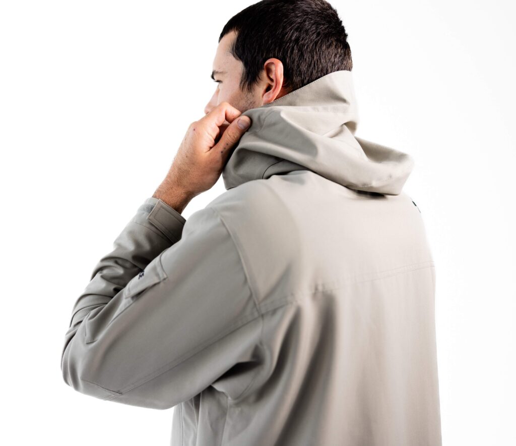 A man wearing a gray jacket with a hood adjusts the collar, facing away against a white background.