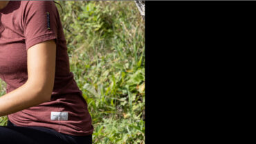 A person wearing a maroon T-shirt sits outdoors, surrounded by green grass, focusing on an object in their hands.