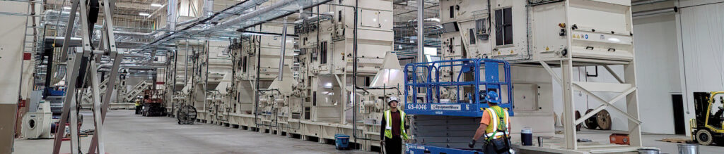 Interior view of a large industrial facility featuring multiple machinery units, with workers in safety gear and equipment in the foreground.