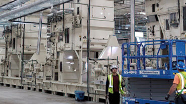 Interior view of a large industrial facility featuring multiple machinery units, with workers in safety gear and equipment in the foreground.