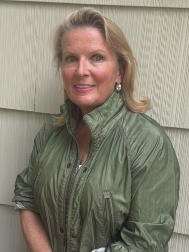 A woman with blonde hair, wearing an olive green jacket, smiles while posing against a beige wall with horizontal panels.