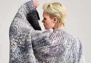 A person with short blonde hair wears a textured cape featuring intricate patterns in shades of gray and blue, partially turned away.