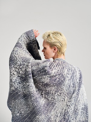 A person with short blonde hair wears a textured cape featuring intricate patterns in shades of gray and blue, partially turned away.