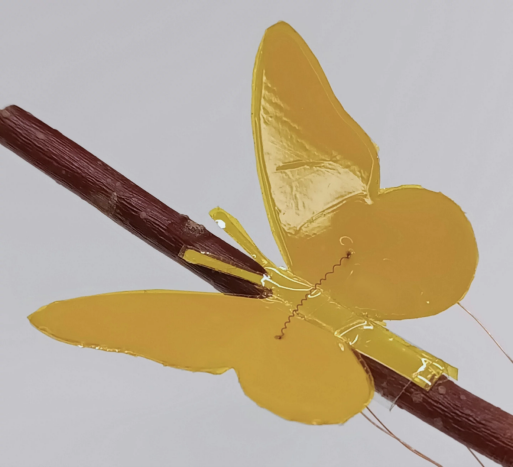 Yellow butterfly-shaped soft robot perched on a brown stick, with delicate wiring visible at the base, against a light background.