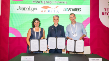 Three individuals stand behind a table holding signed documents, smiling. A colorful backdrop features logos of Jeanologia, HKRITA, and Looptworks.