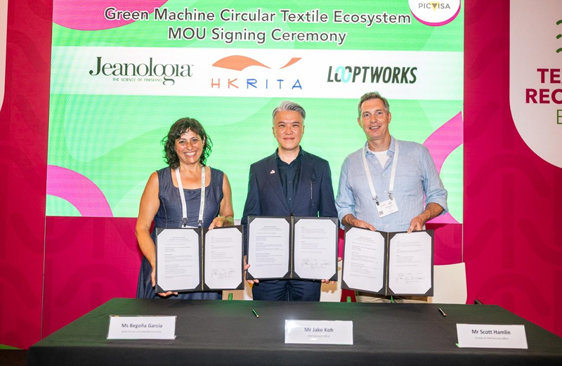 Green Machine Three individuals stand behind a table holding signed documents, smiling. A colorful backdrop features logos of Jeanologia, HKRITA, and Looptworks.