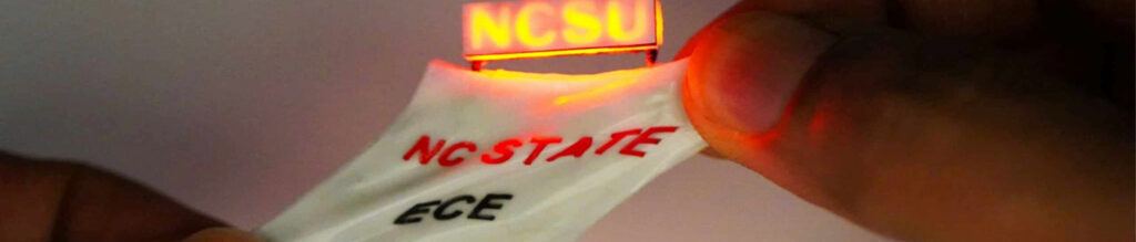 A hand holds a flexible strip with glowing red "NCSU" text, combined with "NC STATE" and "ECE" printed in bold, contrasting colors.