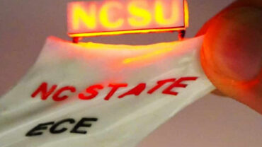 A hand holds a flexible strip with glowing red "NCSU" text, combined with "NC STATE" and "ECE" printed in bold, contrasting colors.