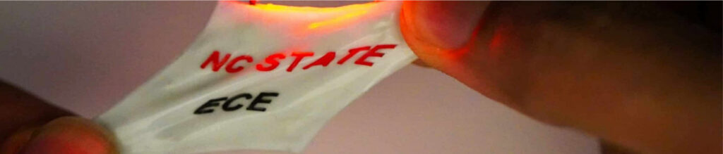 A close-up of a hand holding a flexible, illuminated fabric with "NC STATE" in red and "ECE" in black. The fabric glows with a soft light.